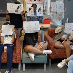 Goup of kids seated in chairs holding pitcures in front of faces. 